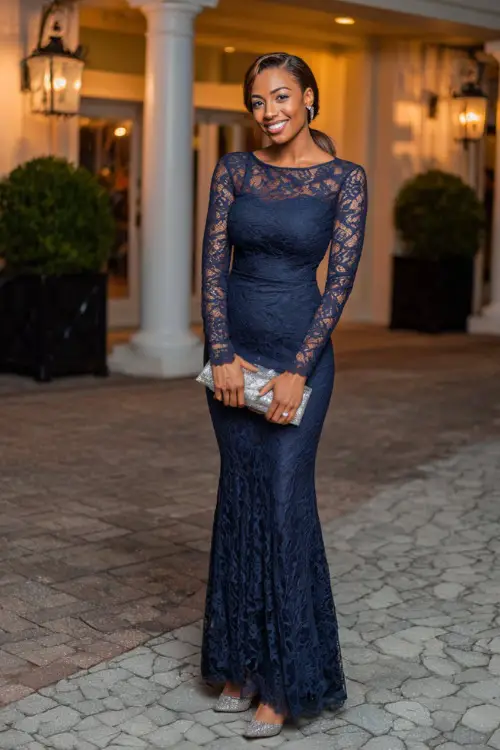 A woman wears a navy blue long-sleeve lace gown with a mermaid silhouette, silver heels, and a metallic clutch at an elegant evening reception 