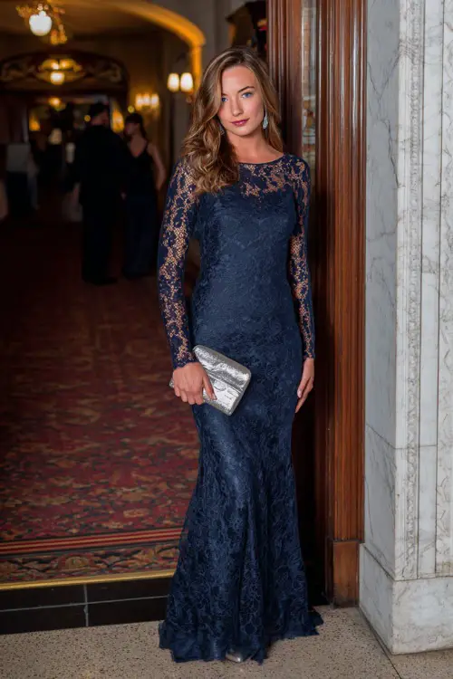 A woman wears a navy blue long-sleeve lace gown with a mermaid silhouette, silver heels, and a metallic clutch at an elegant evening reception