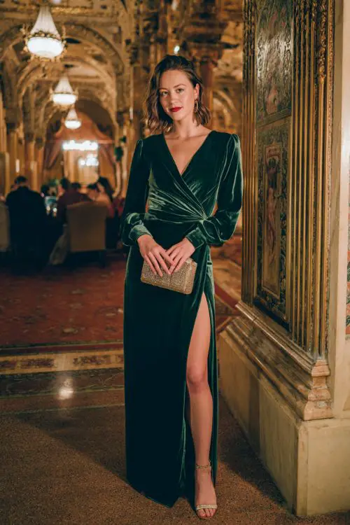 A woman wears an emerald green velvet wrap gown with a thigh-high slit, metallic heels, and a jeweled clutch in a grand historic ballroom 