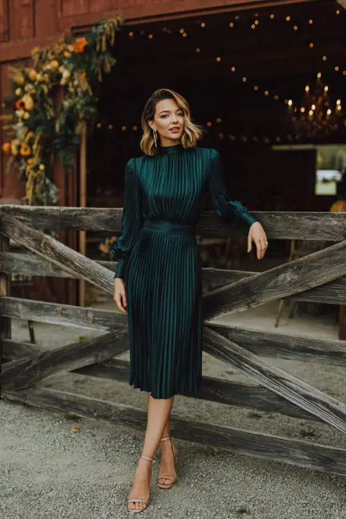 Elegant woman in a dark emerald green pleated midi dress with a high neckline and heeled sandals, posing near a barn decorated for a fall wedding 