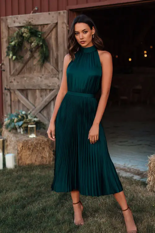 Elegant woman in a dark emerald green pleated midi dress with a high neckline and heeled sandals, posing near a barn decorated for a fall wedding