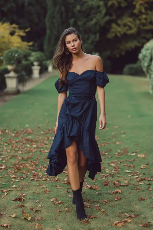 Elegant woman in a navy off-the-shoulder midi dress with ruffled hem and ankle boots, walking across a grassy wedding lawn scattered with leaves 