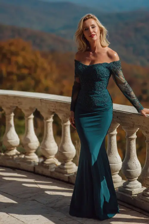 Mature woman in a dark teal mermaid gown with sheer sleeves and subtle beading, posing on a terrace with sweeping views of October trees, full body