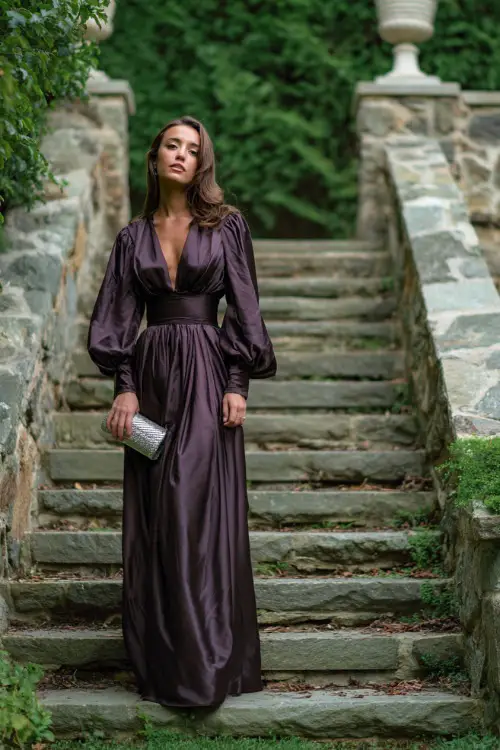 Woman in a black tie-worthy dark mauve satin gown with long bishop sleeves and silver clutch, standing on a leaf-covered stone staircase, full body