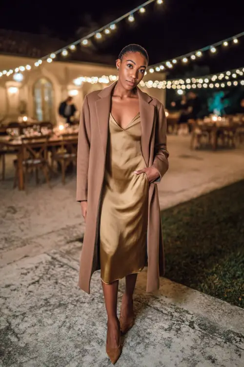 Woman in a bronze satin slip dress with a cowl neckline, paired with a long tailored coat and suede pumps, standing in an outdoor wedding setting with string lights 