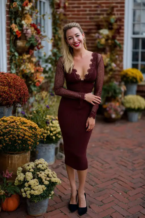 Woman in a burgundy midi sheath dress with lace detailing and black block heels, standing on a brick patio surrounded by fall floral arrangements 