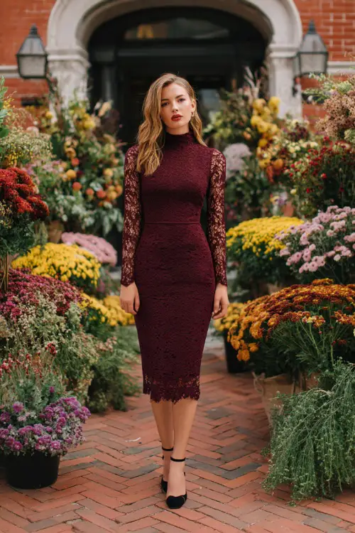 Woman in a burgundy midi sheath dress with lace detailing and black block heels, standing on a brick patio surrounded by fall floral arrangements 