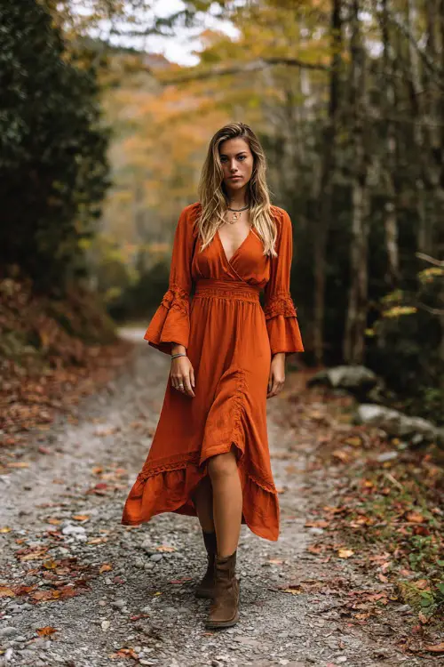 Woman in a burnt orange boho midi dress with flared sleeves and ankle boots, standing in a forest path surrounded by October leaves, full body