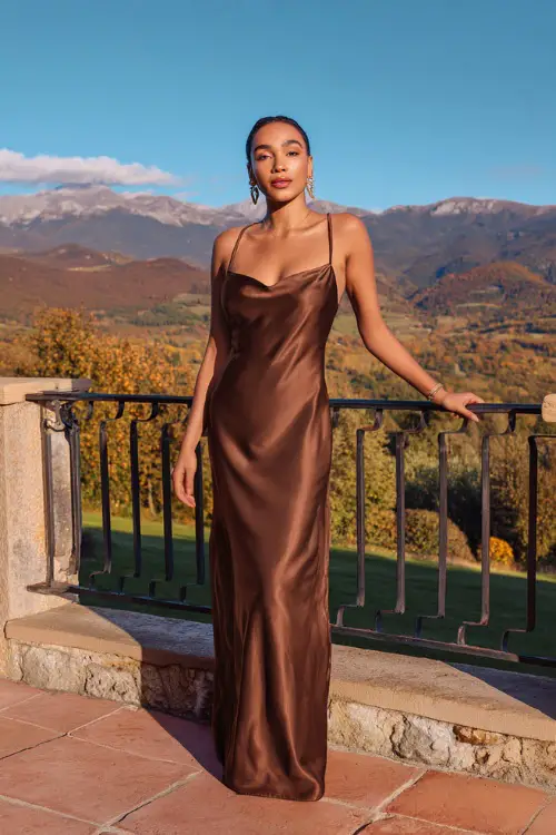 Woman in a chocolate brown satin gown with thin straps and an A-line silhouette, styled with gold hoop earrings and a small metallic clutch, standing on a terrace overlooking a fall landscape