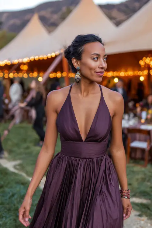 Woman in a dark mauve fit-and-flare dress with pleated skirt and statement earrings, walking near an October wedding reception tent with string lights 