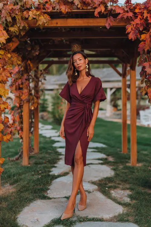Woman in a deep burgundy midi dress with a wrap waist and flutter sleeves, paired with pointed-toe heels and gold earrings