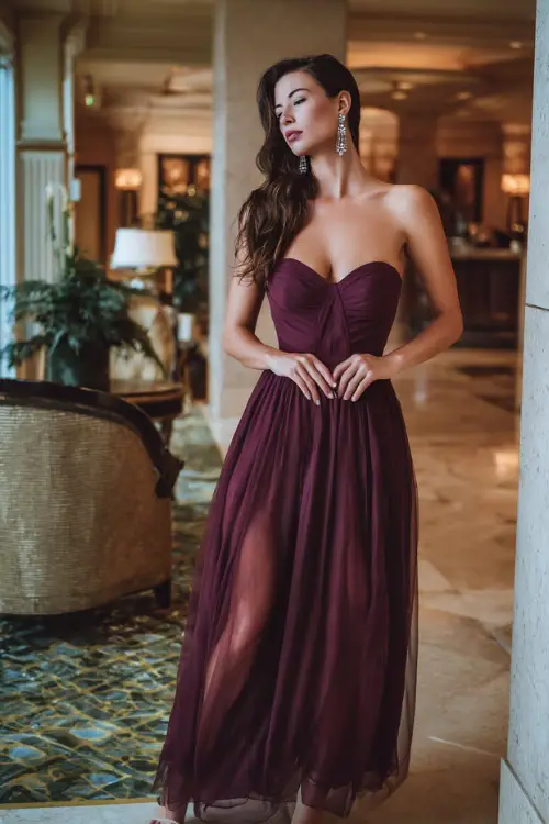 Woman in a deep wine-colored gown with a sweetheart neckline and chiffon overlay, accessorized with diamond drop earrings and heels, posing in a luxurious hotel lobby 