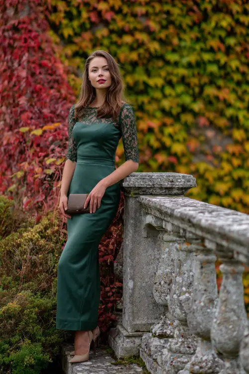 Woman in a forest green column dress with lace sleeves and satin clutch, standing by stone balustrades with ivy turning autumn red