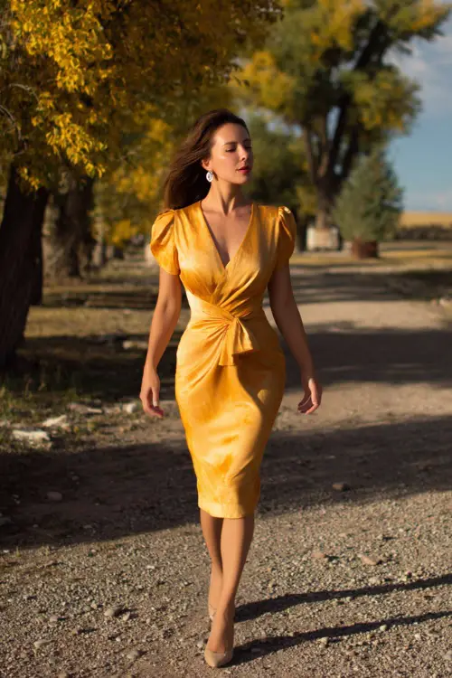 Woman in a goldenrod wrap midi dress with subtle shimmer, paired with low heels and pearl earrings, walking on gravel under golden trees, full body