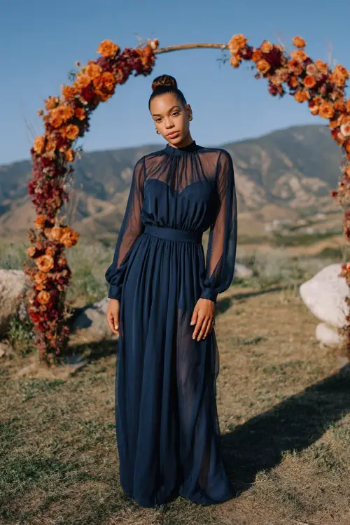 Woman in a navy chiffon maxi dress with sheer long sleeves and a cinched waist, standing near a ceremony arch decorated with orange and burgundy flowers 