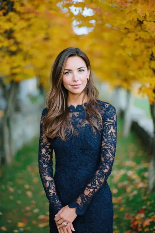 Woman in a navy lace dress with scalloped hem and long sleeves, posing under golden maple trees at a scenic October wedding venue, full body 