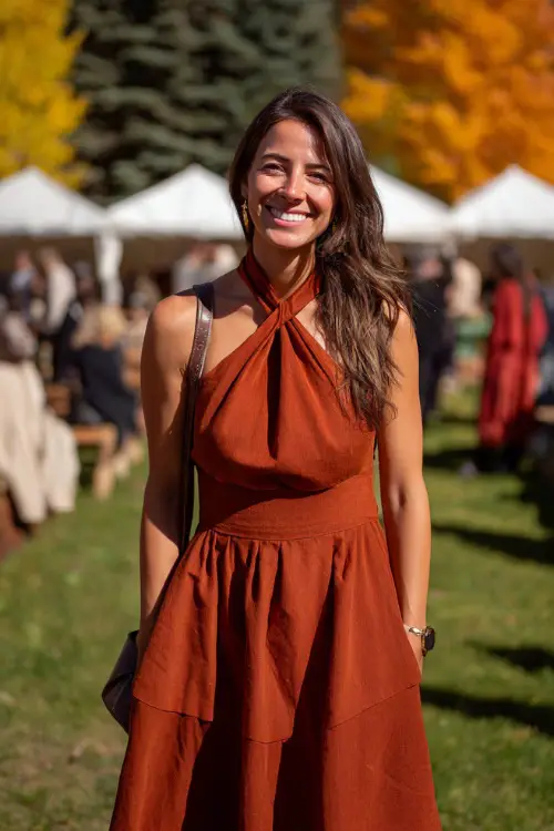 Woman in a rust-colored halter midi dress with layered skirt and crossbody bag, smiling at an open-air ceremony with autumn trees in the background 