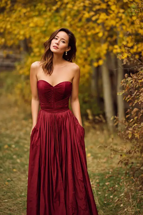 Woman over 40 wearing a floor-length burgundy gown with a pleated skirt and structured bodice, posing near golden autumn trees at an outdoor October wedding