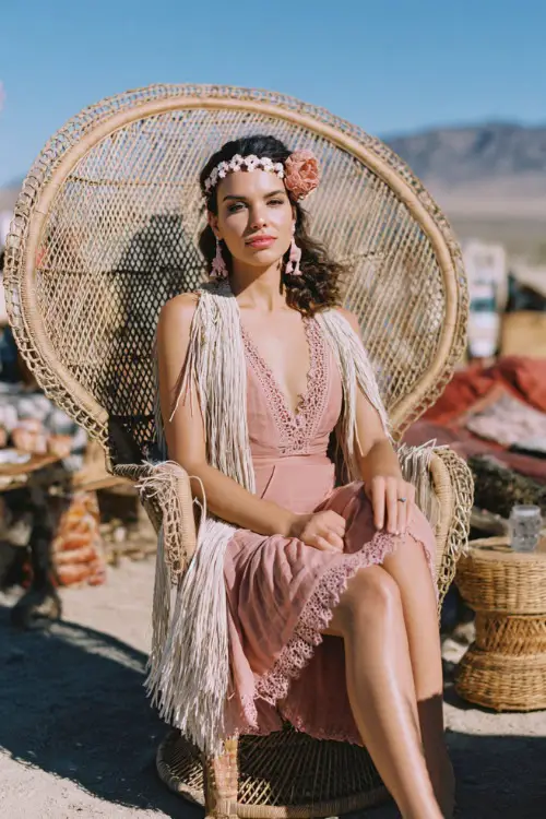 Woman wearing a blush pink crochet-trimmed midi dress with fringe shawl, sitting on a rattan chair at a desert-inspired October wedding, full body 