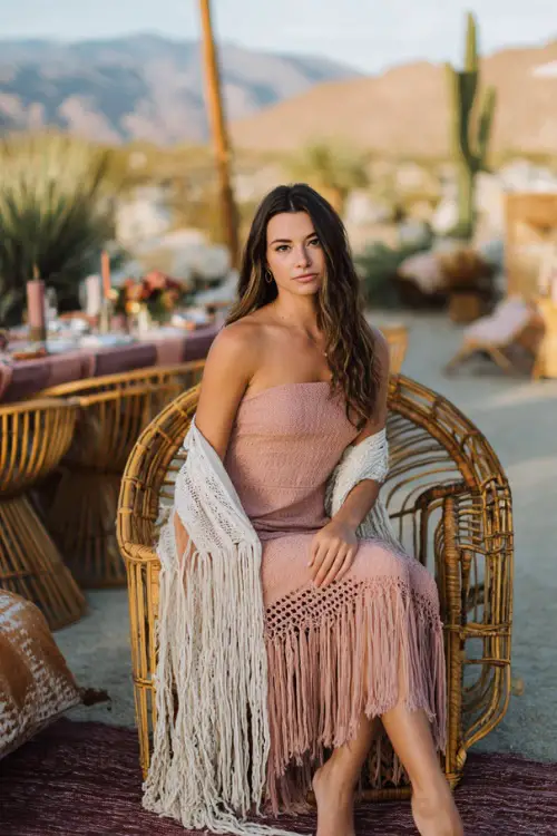 Woman wearing a blush pink crochet-trimmed midi dress with fringe shawl, sitting on a rattan chair at a desert-inspired October wedding, full body