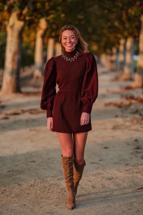 Woman wearing a burgundy puff sleeve mini dress with a high neckline, paired with knee-high suede boots and a statement necklace