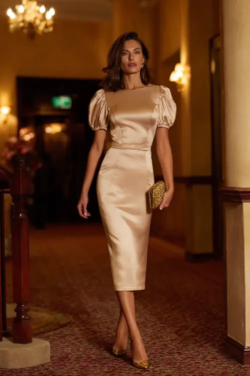 Woman wearing a champagne satin tea-length dress with short puff sleeves and a fitted waist, paired with gold heels and a matching clutch 