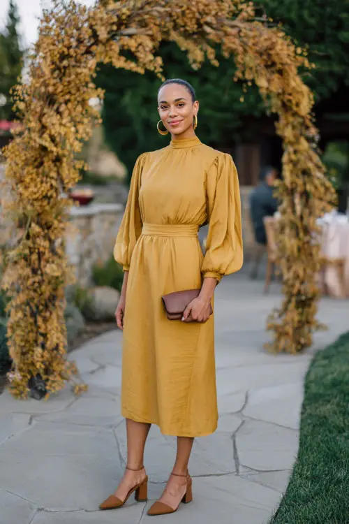 Woman wearing a mustard yellow midi dress with bishop sleeves and a high neckline, paired with brown block heels and a soft leather clutch