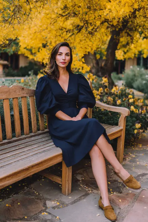 Woman wearing a navy A-line midi dress with puff sleeves and suede loafers, seated on a wooden bench at a casual fall wedding venue surrounded by yellow foliage 