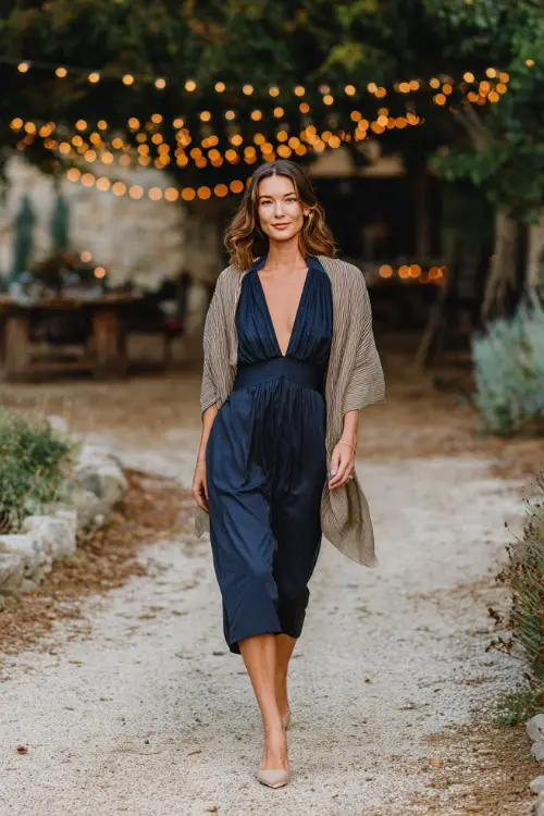 Woman wearing a navy dress with pleated skirt and keyhole neckline, styled with beige heels and a soft shawl, walking under a string-light canopy in a rustic October wedding setting 