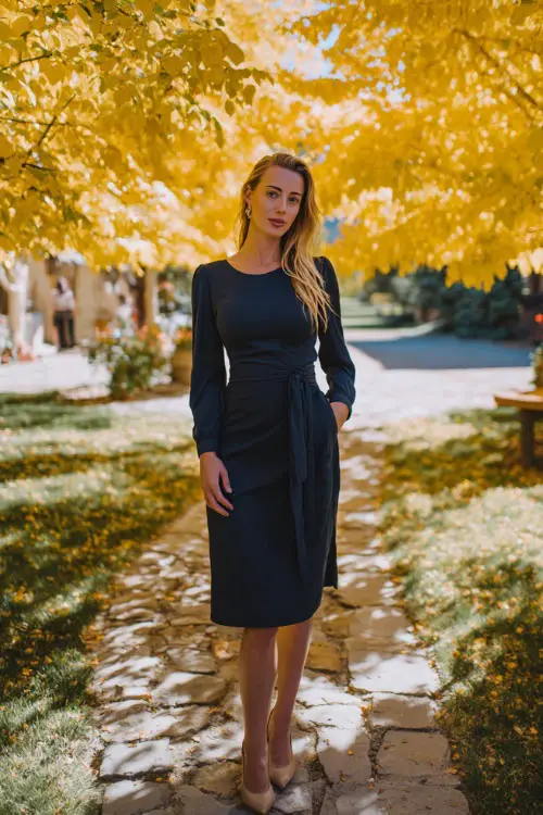 Woman wearing a navy midi dress with long sleeves and a tie waist, standing beneath golden autumn trees at an outdoor October wedding venue