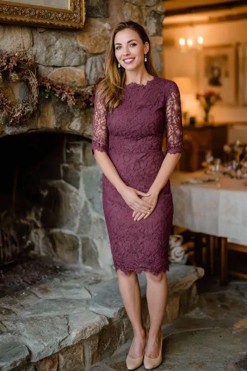 Woman wearing a plum lace sheath dress with scalloped edges and three-quarter sleeves, accessorized with pearl earrings and beige pumps 