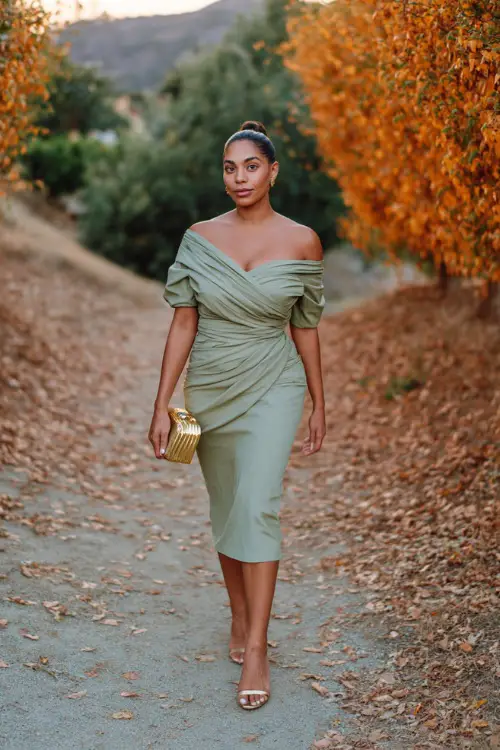 Woman wearing a sage green off-the-shoulder midi dress with ruched detail, styled with gold heels and a small clutch