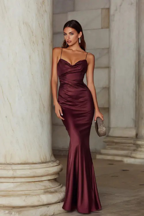 Woman wearing a sleek wine satin gown with thin straps and a mermaid hemline, accessorized with diamond studs and a small metallic clutch, standing by a marble column 