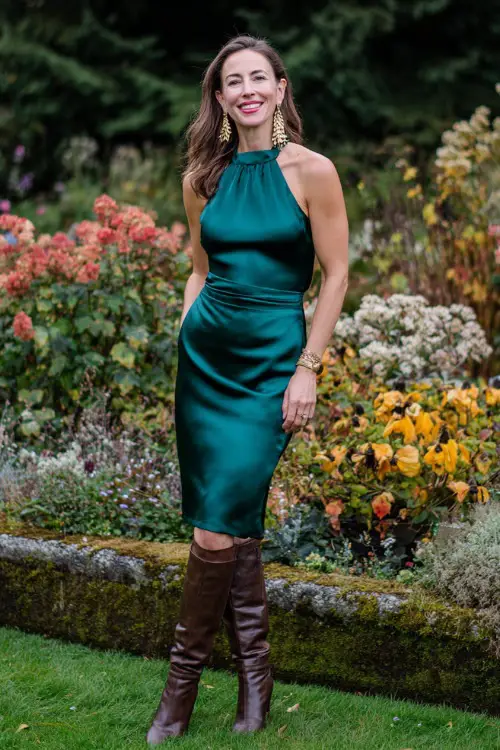 chic fall wedding guest outfit, midi satin dress in rich emerald green, paired with knee-high leather boots, gold drop earrings, autumn garden backdrop
