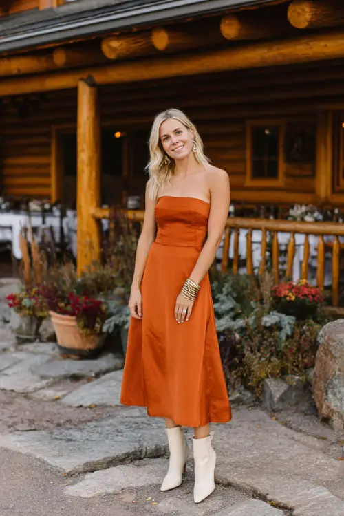 elegant autumn wedding guest look, burnt orange A-line midi dress, cream ankle boots, layered gold bracelets, mountain lodge wedding backdrop