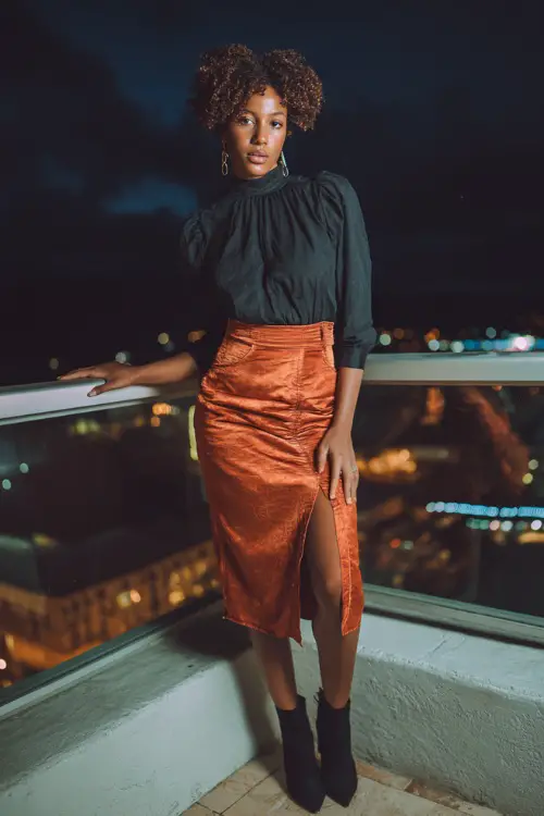 A Black woman in a chic burnt orange slip skirt paired with a black fitted blouse and ankle boots, standing on a rooftop terrace at night, full body