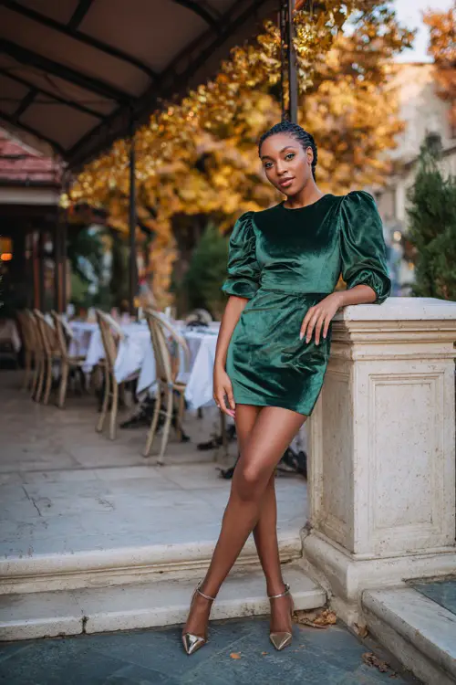 A Black woman in a chic green velvet mini dress with puff sleeves and metallic heels, posing near a fall café decorated with golden leaves, full body