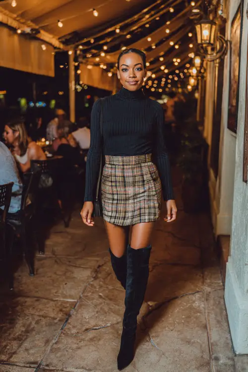 A Black woman in a chic plaid mini skirt with a fitted turtleneck and over-the-knee boots, walking into a lively fall dinner spot