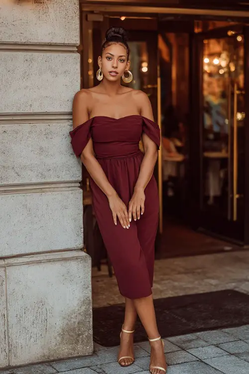 A beautiful Black woman in a burgundy off-shoulder midi dress with gold heels and hoop earrings, standing in front of a candlelit restaurant entrance