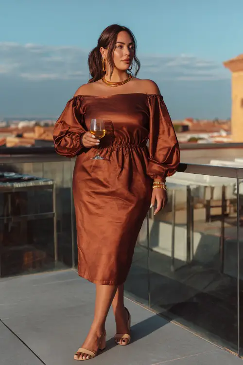 A plus-size woman in a copper satin midi dress with puff sleeves, styled with gold jewelry and block heels, holding a glass of wine at a rooftop dinner terrace in the fall