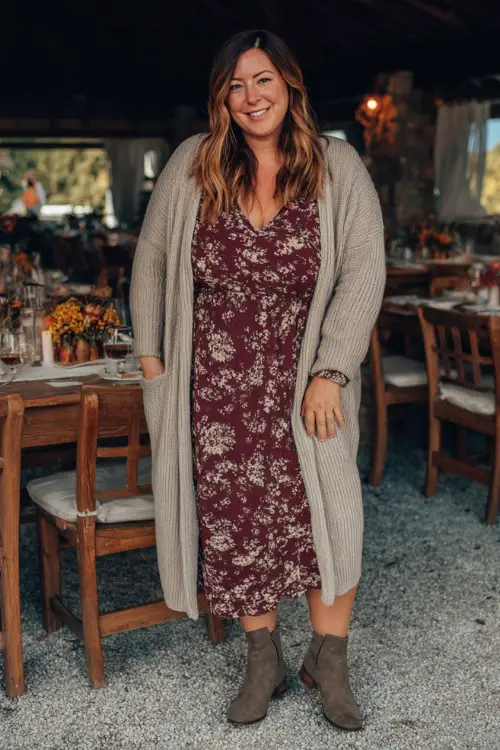 A plus-size woman in a long cardigan layered over a floral midi dress with ankle boots, smiling at a cozy outdoor fall dinner spot