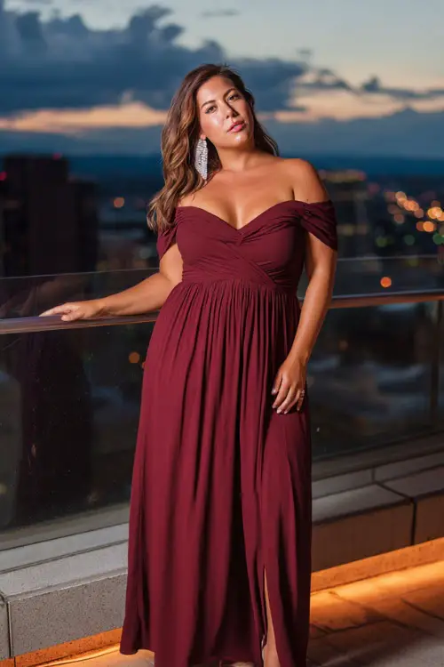 A plus-size woman in a maroon off-the-shoulder maxi dress paired with silver earrings and block heels, standing on a glowing rooftop terrace at night 