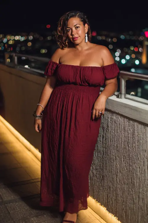 A plus-size woman in a maroon off-the-shoulder maxi dress paired with silver earrings and block heels, standing on a glowing rooftop terrace at night 