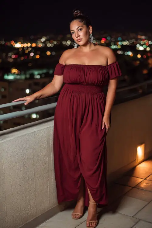 A plus-size woman in a maroon off-the-shoulder maxi dress paired with silver earrings and block heels, standing on a glowing rooftop terrace at night