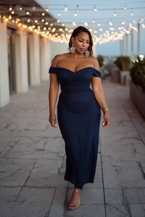 A plus-size woman in a navy blue off-shoulder maxi dress with silver heels and chandelier earrings, walking through a rooftop terrace glowing with fall string lights 