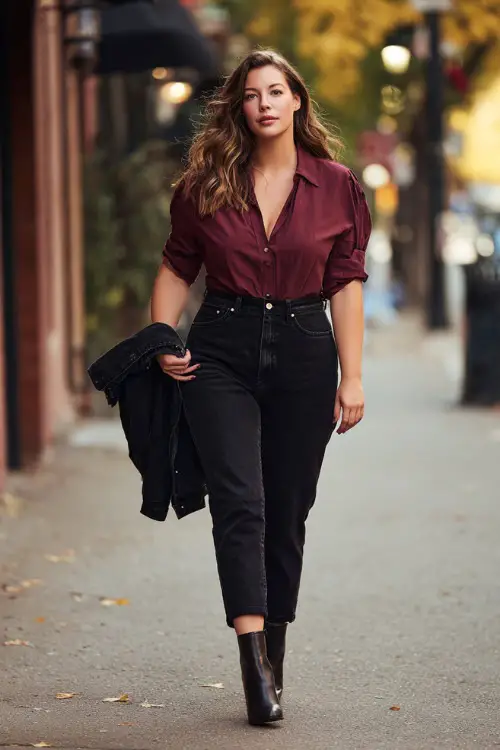 A plus-size woman in black high-waisted jeans, a burgundy blouse, and block-heeled ankle boots, holding a jacket while walking down an autumn street