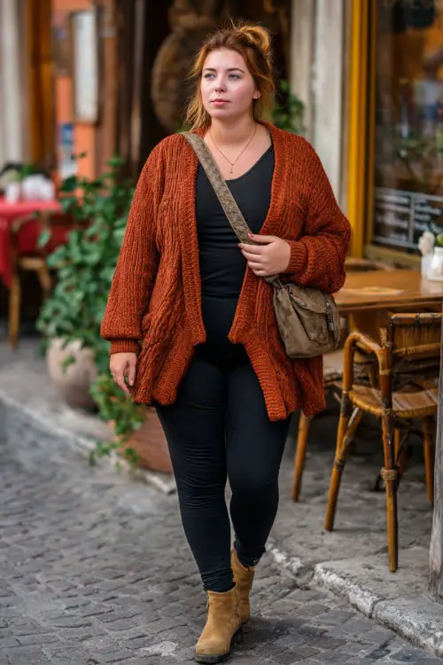 A plus-size woman in black skinny jeans, a rust-colored knit cardigan, and suede ankle boots, holding a crossbody bag while strolling past a casual fall restaurant