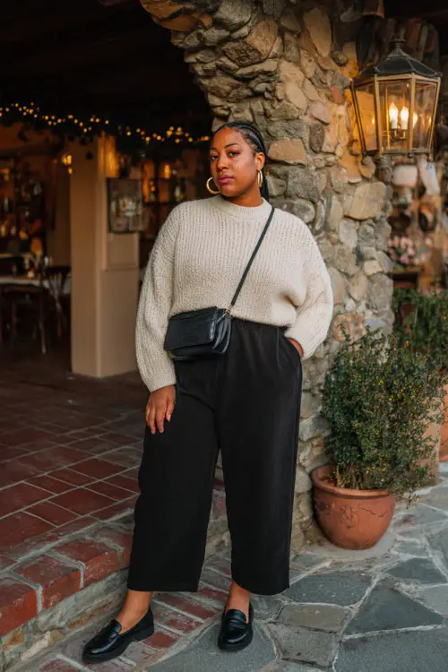 A plus-size woman in black wide-leg jeans, a cream knit sweater tucked in, accessorized with hoop earrings and loafers