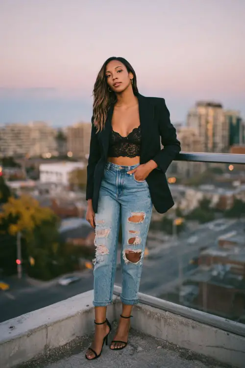A slim woman in ripped straight-leg jeans with a black blazer layered over a lace camisole, styled with stiletto heels, standing on an autumn city rooftop at dusk