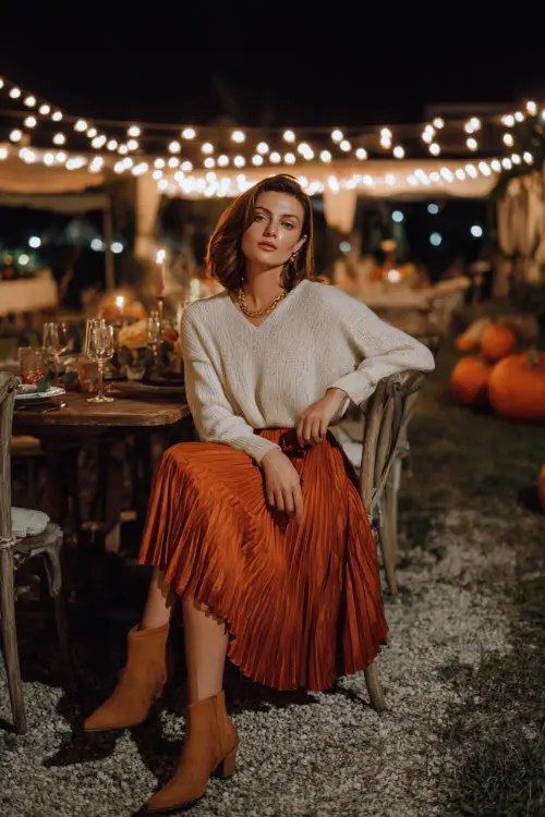 A woman in a burnt orange pleated midi skirt with a cream sweater and heeled booties, dining outdoors under string lights with pumpkins around 